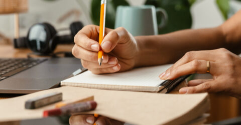 Mains d'une femme qui tient un crayon et un carnet devant un ordinateur.